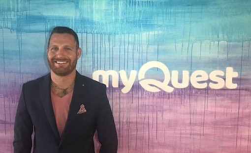 Rob Matzkin with blue jacket posing in front of wall with myQuest logo